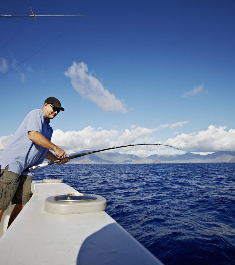 Mature man reeling in fish on deck of sport fishing boat island of Oahu in background